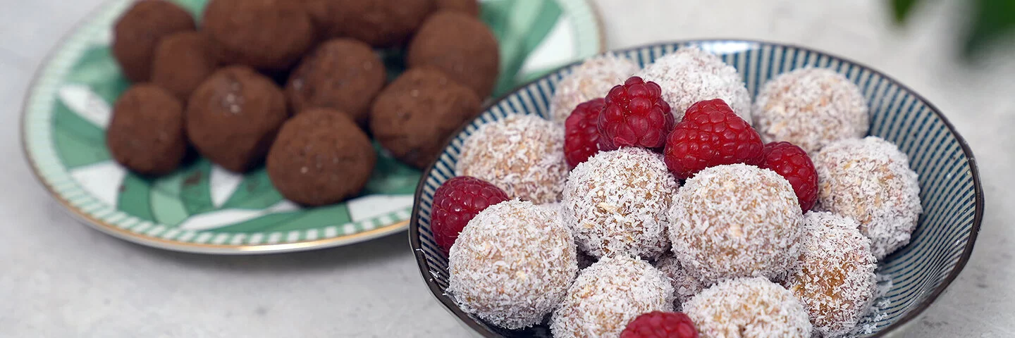Im Vordergrund Kokos-Himbeer Energy Balls nach einem Rezept von Felicitas Then, angerichtet auf einem schwarz-weiß gestreiften Teller, garniert mit Himbeeren. Im Hintergrund Schoko-Erdnuss Energy Balls auf einem grün-weißem Teller.