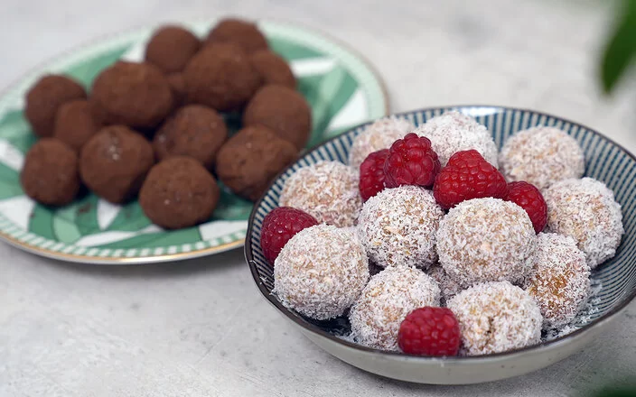 Im Vordergrund Kokos-Himbeer Energy Balls nach einem Rezept von Felicitas Then, angerichtet auf einem schwarz-weiß gestreiften Teller, garniert mit Himbeeren. Im Hintergrund Schoko-Erdnuss Energy Balls auf einem grün-weißem Teller.