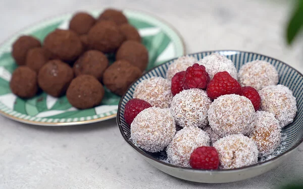 Im Vordergrund Kokos-Himbeer Energy Balls nach einem Rezept von Felicitas Then, angerichtet auf einem schwarz-weiß gestreiften Teller, garniert mit Himbeeren. Im Hintergrund Schoko-Erdnuss Energy Balls auf einem grün-weißem Teller.