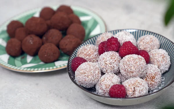 Im Vordergrund Kokos-Himbeer Energy Balls nach einem Rezept von Felicitas Then, angerichtet auf einem schwarz-weiß gestreiften Teller, garniert mit Himbeeren. Im Hintergrund Schoko-Erdnuss Energy Balls auf einem grün-weißem Teller.