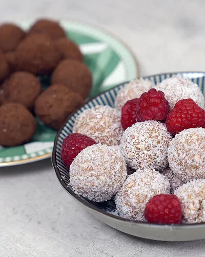 Im Vordergrund Kokos-Himbeer Energy Balls nach einem Rezept von Felicitas Then, angerichtet auf einem schwarz-weiß gestreiften Teller, garniert mit Himbeeren. Im Hintergrund Schoko-Erdnuss Energy Balls auf einem grün-weißem Teller.