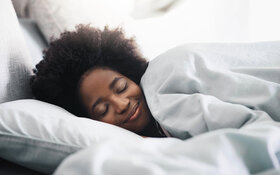 Eine junge Frau liegt mit geschlossenen Augen seitlich im Bett und lächelt.