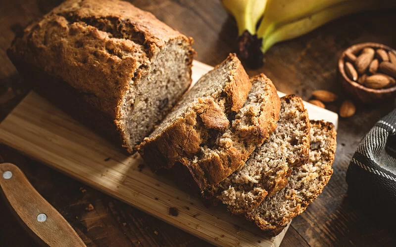 Kastenförmiges Bananenbrot liegt angeschnitten auf einem Holzbrett auf Holzuntergrund.