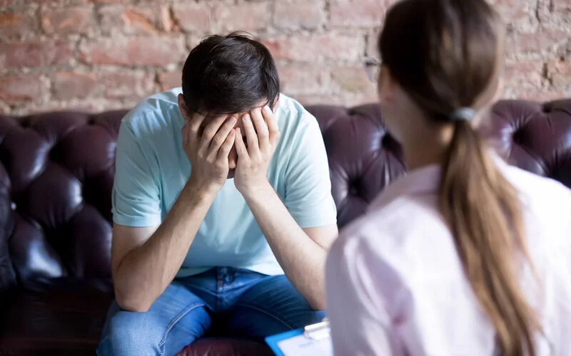 Ein junger Mann sitzt gegenüber seiner Therapeutin auf einem Sofa und hat die Hände vor das Gesicht geschlagen.