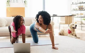 Mutter und Tochter machen Yoga im Wohnzimmer. Vor ihnen steht ein aufgeklappter Laptop, im Hintergrund sind eine helle Wohnzimmereinrichtung und ein Golden Retriever zu sehen.