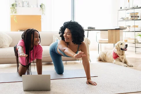 Mutter und Tochter machen Yoga im Wohnzimmer. Vor ihnen steht ein aufgeklappter Laptop, im Hintergrund sind eine helle Wohnzimmereinrichtung und ein Golden Retriever zu sehen.