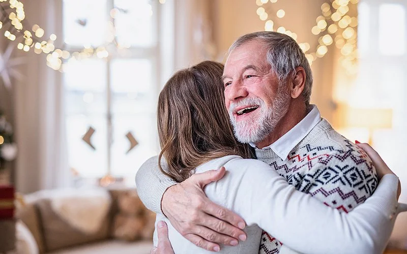 Eine jüngere Frau, die von hinten zu sehen ist, umarmt einen älteren Mann, im Hintergrund Weihnachtsdekoration und eine Lichterkette.