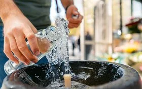 Hände halten eine Glasflasche unter einen Quellstrahl; Wasser spritzt in ein rundes Steinbecken.