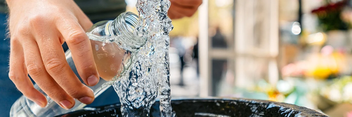 Hände halten eine Glasflasche unter einen Quellstrahl; Wasser spritzt in ein rundes Steinbecken.