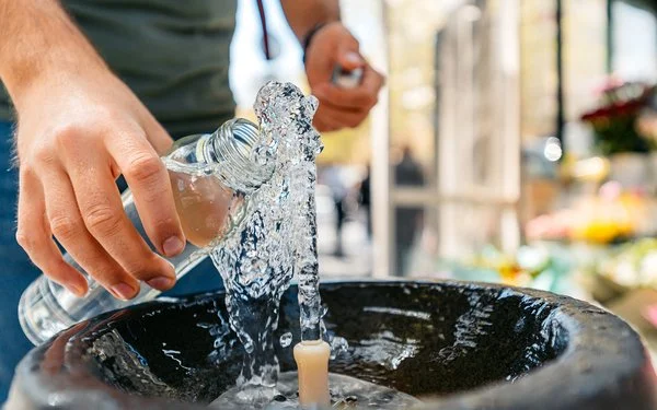 Hände halten eine Glasflasche unter einen Quellstrahl; Wasser spritzt in ein rundes Steinbecken.