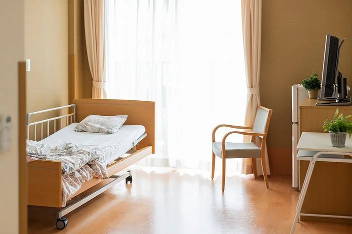 Pflegezimmer mit einem Pflegebett. Gardinen, Vorhänge, Topfpflanzen und ein Fernseher sorgen für eine gemütliche Atmosphäre.