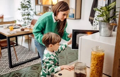 Eine Mutter und ihr kleiner Sohn sind seitlich zu sehen. Sie öffnen die Tür einer Mikrowelle und schauen hinein.