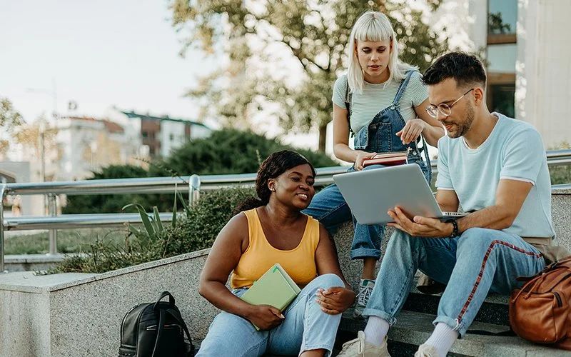 Zwei jüngere Frauen und ein Mann sitzen auf einer Treppe und schauen auf einen Laptop.
