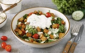 Weiße Schale mit knusprigen Kartoffelecken und selbstgemachtem Tsatsiki auf grünem Salat mit Fetawürfeln und Tomaten auf einem grauen Untergrund. Rechts daneben Messer und Gabel. Im Hintergrund Tomaten, Gurke und Lollo Rosso.