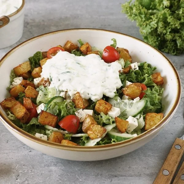 Weiße Schale mit knusprigen Kartoffelecken und selbstgemachtem Tsatsiki auf grünem Salat mit Fetawürfeln und Tomaten auf einem grauen Untergrund. Rechts daneben Messer und Gabel. Im Hintergrund Tomaten, Gurke und Lollo Rosso.