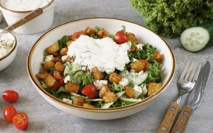 Weiße Schale mit knusprigen Kartoffelecken und selbstgemachtem Tsatsiki auf grünem Salat mit Fetawürfeln und Tomaten auf einem grauen Untergrund. Rechts daneben Messer und Gabel. Im Hintergrund Tomaten, Gurke und Lollo Rosso.