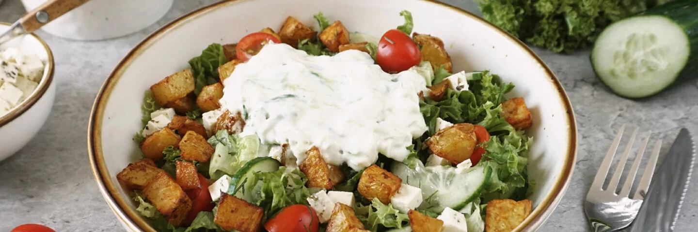 Weiße Schale mit knusprigen Kartoffelecken und selbstgemachtem Tsatsiki auf grünem Salat mit Fetawürfeln und Tomaten auf einem grauen Untergrund. Rechts daneben Messer und Gabel. Im Hintergrund Tomaten, Gurke und Lollo Rosso.