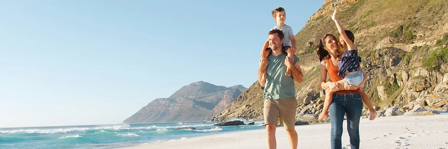 Eine Familie mit zwei Erwachsenen und zwei Kindern läuft am Strand entlang. Ein Erwachsener trägt ein Kind auf den Schultern, während das andere Kind von der anderen erwachsenen Person getragen wird. Im Hintergrund sind das Meer, Wellen und felsige Hügel zu sehen. Der Himmel ist klar und blau.