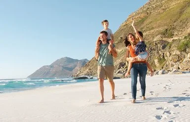 Eine Familie mit zwei Erwachsenen und zwei Kindern läuft am Strand entlang. Ein Erwachsener trägt ein Kind auf den Schultern, während das andere Kind von der anderen erwachsenen Person getragen wird. Im Hintergrund sind das Meer, Wellen und felsige Hügel zu sehen. Der Himmel ist klar und blau.