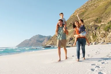 Eine Familie mit zwei Erwachsenen und zwei Kindern läuft am Strand entlang. Ein Erwachsener trägt ein Kind auf den Schultern, während das andere Kind von der anderen erwachsenen Person getragen wird. Im Hintergrund sind das Meer, Wellen und felsige Hügel zu sehen. Der Himmel ist klar und blau.