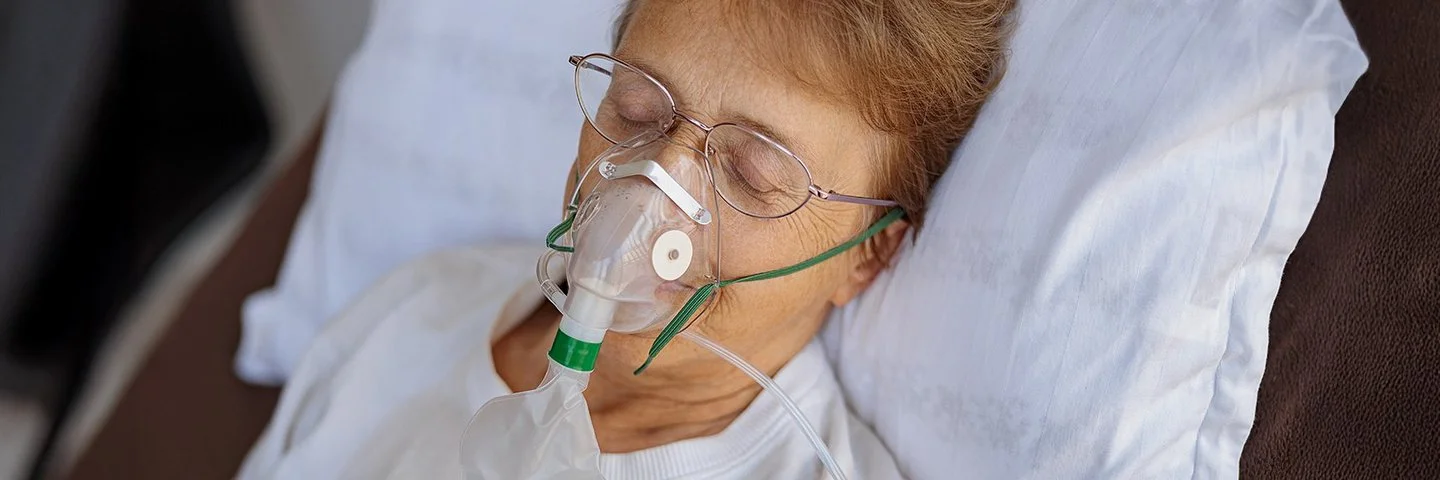 Eine ältere Frau mit Brille liegt mit geschlossenen Augen in einem Bett und trägt eine Sauerstoffmaske.