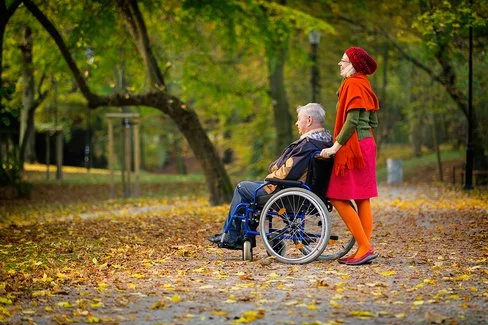 Eine Frau schiebt einen älteren Mann im Rollstuhl durch einen herbstlichen Park. Der Boden ist mit bunten Blättern bedeckt, die Bäume tragen gelb-grünes Laub. Die Frau trägt einen roten Rock, eine orangefarbene Strickjacke und eine rote Mütze, der Mann trägt dunkle Kleidung.