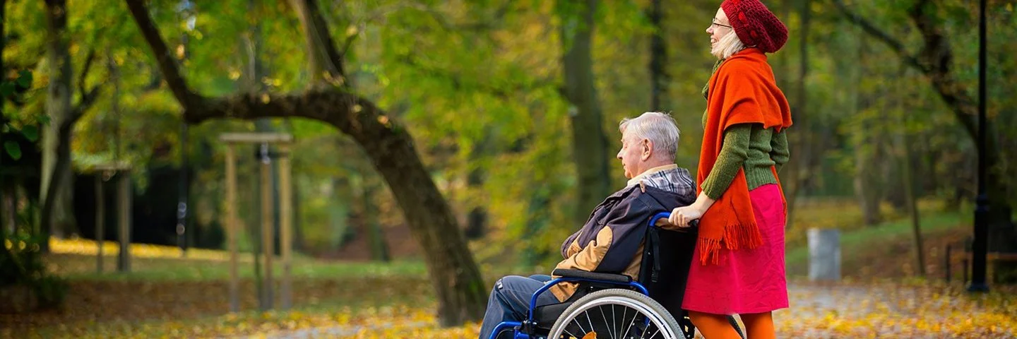 Eine Frau schiebt einen älteren Mann im Rollstuhl durch einen herbstlichen Park. Der Boden ist mit bunten Blättern bedeckt, die Bäume tragen gelb-grünes Laub. Die Frau trägt einen roten Rock, eine orangefarbene Strickjacke und eine rote Mütze, der Mann trägt dunkle Kleidung.
