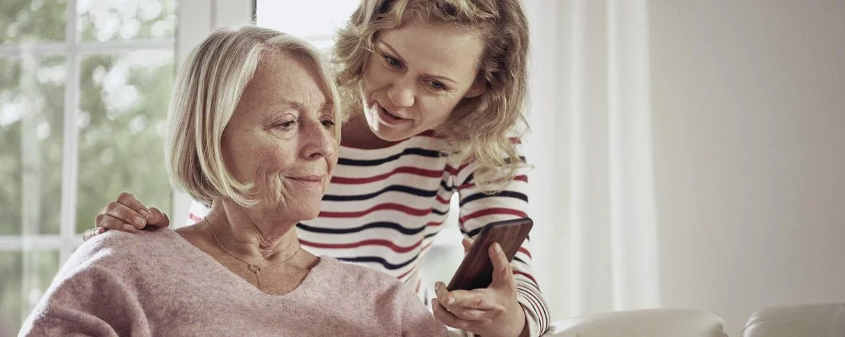 Eine ältere Frau sitzt auf einem Sofa und nutzt ein Smartphone, während eine jüngere Frau hinter dem Sofa steht und über die Schulter blickt, um etwas auf ihrem Smartphone zu zeigen.