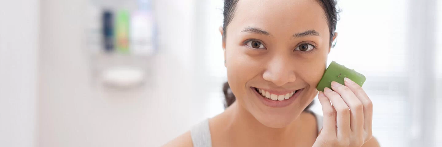 Eine junge Frau steht im Bad und reibt sich über die Gesichtshaut mit einem frisch aufgeschnitten Blatt Aloe vera.