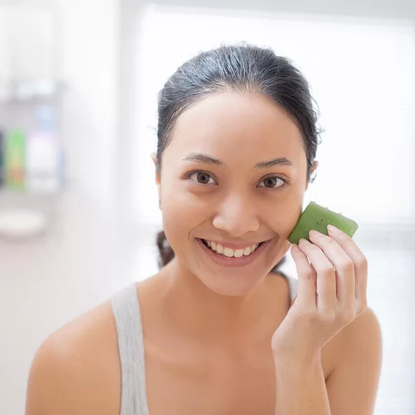 Eine junge Frau steht im Bad und reibt sich über die Gesichtshaut mit einem frisch aufgeschnitten Blatt Aloe vera.