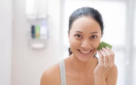 Eine junge Frau steht im Bad und reibt sich über die Gesichtshaut mit einem frisch aufgeschnitten Blatt Aloe vera.