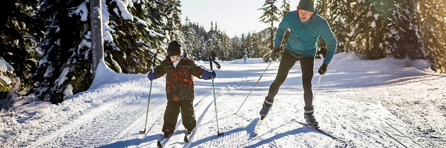Erwachsener und Kind beim Langlaufen auf einer präparierten Loipe im Winterwald. Das Kind fährt in der klassischen Spur, der Erwachsene skatet daneben und unterstützt es.