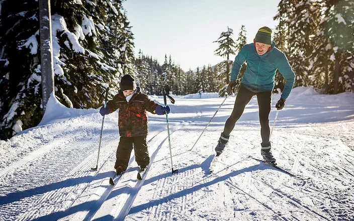 Erwachsener und Kind beim Langlaufen auf einer präparierten Loipe im Winterwald. Das Kind fährt in der klassischen Spur, der Erwachsene skatet daneben und unterstützt es.