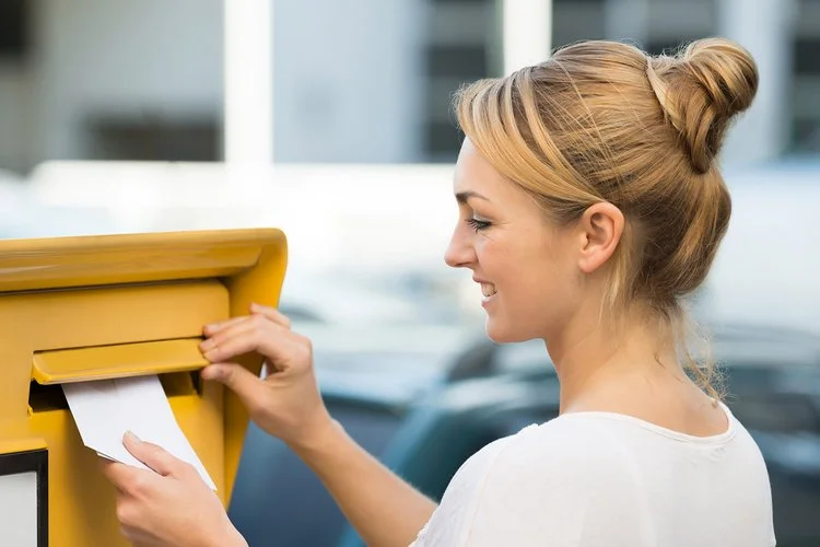 Eine junge Frau wirft lächelnd Briefe in einen Postbriefkasten.