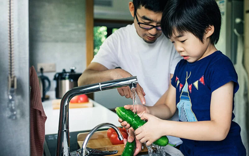 Ein Vater wäscht mit seinem Sohn eine Salatgurke über dem Waschbecken, um mögliche Krankheitserreger zu entfernen.