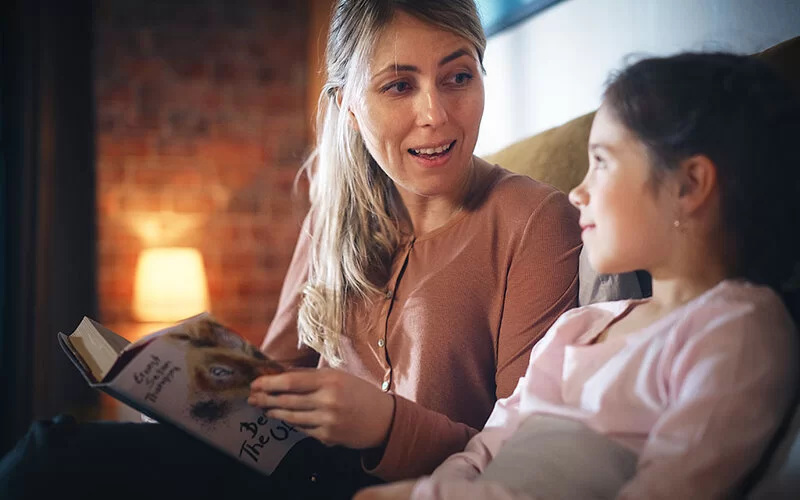 Eine Mutter und ihre Tochter sitzen bei dämmrigem Licht im Schlafzimmer auf dem Bett. Die Mutter liest eine Geschichte aus einem Buch vor, die Tochter lächelt sie an.