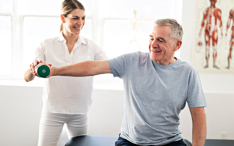 Eine Physiotherapeutin zeigt einem Senioren, wie er eine Gymnastikübung mit einer Hantel im Sitzen ausführen kann.