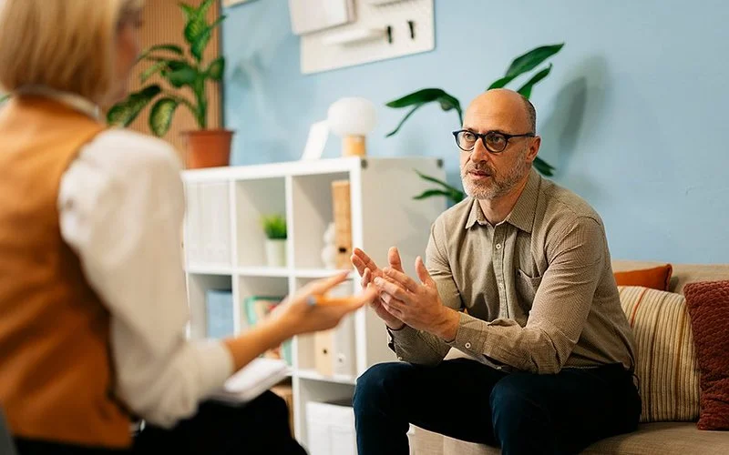 Ein Mann mit Brille und Bart sitzt auf einem Sofa in einem modern eingerichteten Raum. Er trägt ein beiges Hemd und dunkle Hose. Im Hintergrund stehen Pflanzen und ein Regal mit verschiedenen Gegenständen. Gegenüber sitzt eine Person mit blonden Haaren, die eine Weste über einem weißen Oberteil trägt und einen Notizblock hält. Die Szene wirkt wie ein Gespräch oder eine Beratungssituation.
