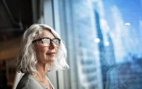 Ältere Frau mit Brille schaut aus einem Fenster in die Ferne.