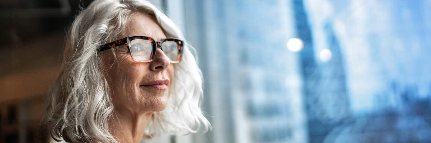 Ältere Frau mit Brille schaut aus einem Fenster in die Ferne.
