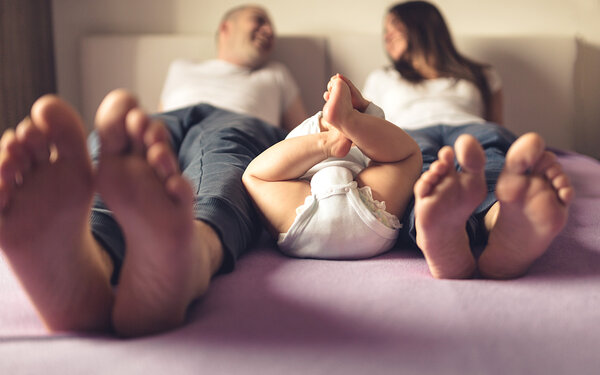 Eine junge Familie mit Baby liegt auf dem Bett, die Füße Richtung Kamera gestreckt.