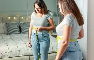 Eine Frau blickt kritisch auf das Maßband, das sie sich um die Talie gelegt hat.