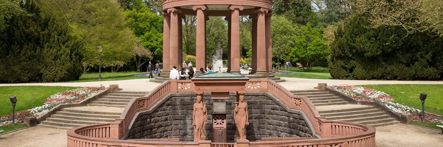 Rundpavillon mit Säulen und Trinkbrunnen in einer Parkanlage, dem Kurpark in Bad Homburg; Menschen spazieren auf den Wegen, Bäume und Beete im Hintergrund.