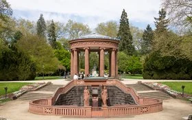 Rundpavillon mit Säulen und Trinkbrunnen in einer Parkanlage, dem Kurpark in Bad Homburg; Menschen spazieren auf den Wegen, Bäume und Beete im Hintergrund.