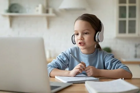 Ein kleines Mädchen sitzt mit Kopfhörern auf den Ohren und einem Filzstift in der Hand vor einem Laptop und schaut auf dessen Bildschirm.