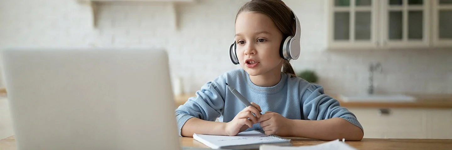 Ein kleines Mädchen sitzt mit Kopfhörern auf den Ohren und einem Filzstift in der Hand vor einem Laptop und schaut auf dessen Bildschirm.