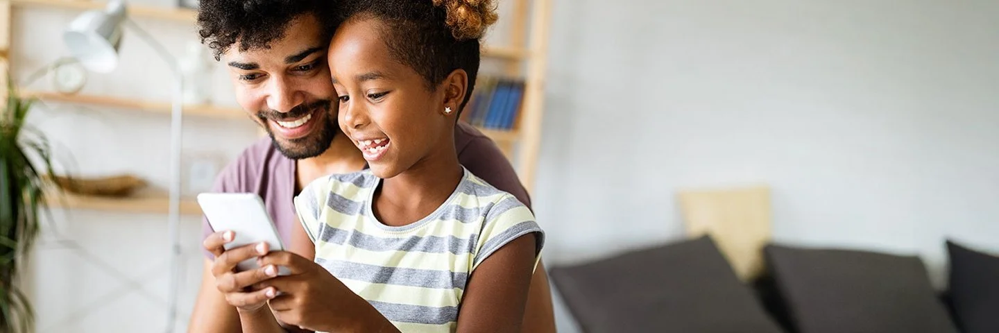 Ein Kind sitzt auf dem Schoß des Vaters auf einem Sofa und hält ein Smartphone in den Händen.