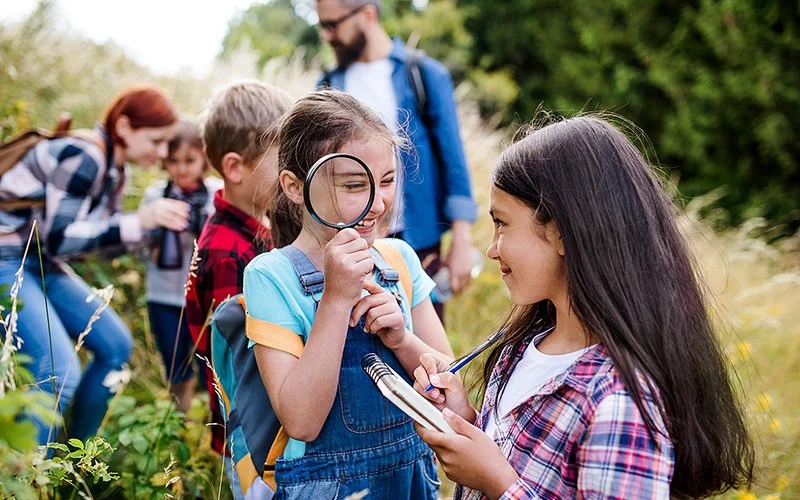 Eine Gruppe von Schulkindern ist mit den Lehrkräften auf Exkursion in der Natur. Das lachende Mädchen im Vordergrund hält eine große Lupe vor ihr Auge. Ein weiteres Mädchen hält einen Block und Stift in der Hand.