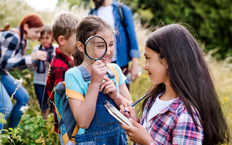 Eine Gruppe von Schulkindern ist mit den Lehrkräften auf Exkursion in der Natur. Das lachende Mädchen im Vordergrund hält eine große Lupe vor ihr Auge. Ein weiteres Mädchen hält einen Block und Stift in der Hand.