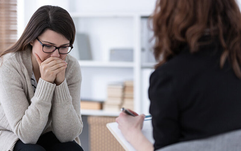 Eine junge Frau sitzt einer Therapeutin in einem Gespräch gegenüber und stützt ihren Kopf mit ihren Händen ab.